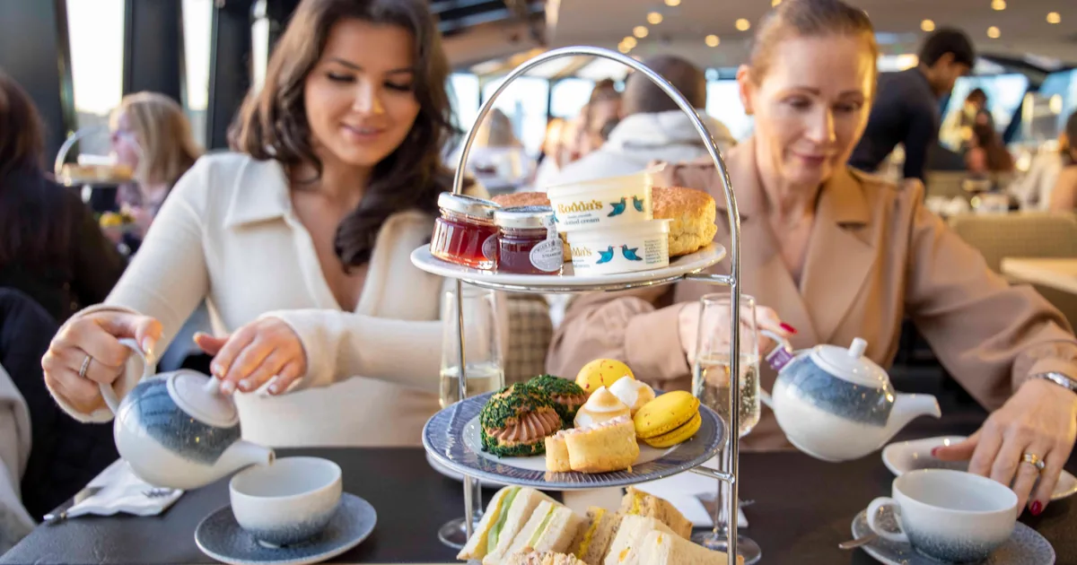 Two guests pouring tea beside a three-tier afternoon tea stand on a Thames cruise boat