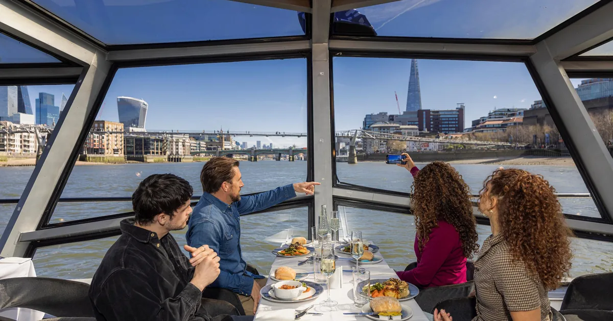 Guests at lunch on a glass-roofed Thames cruise boat, one pointing out the Shard