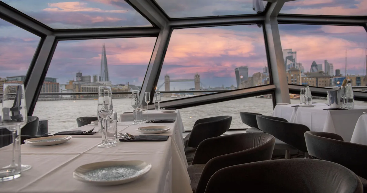 The glass-walled dining deck of a Thames cruise boat with laid tables, the Shard and Tower Bridge outside at dusk